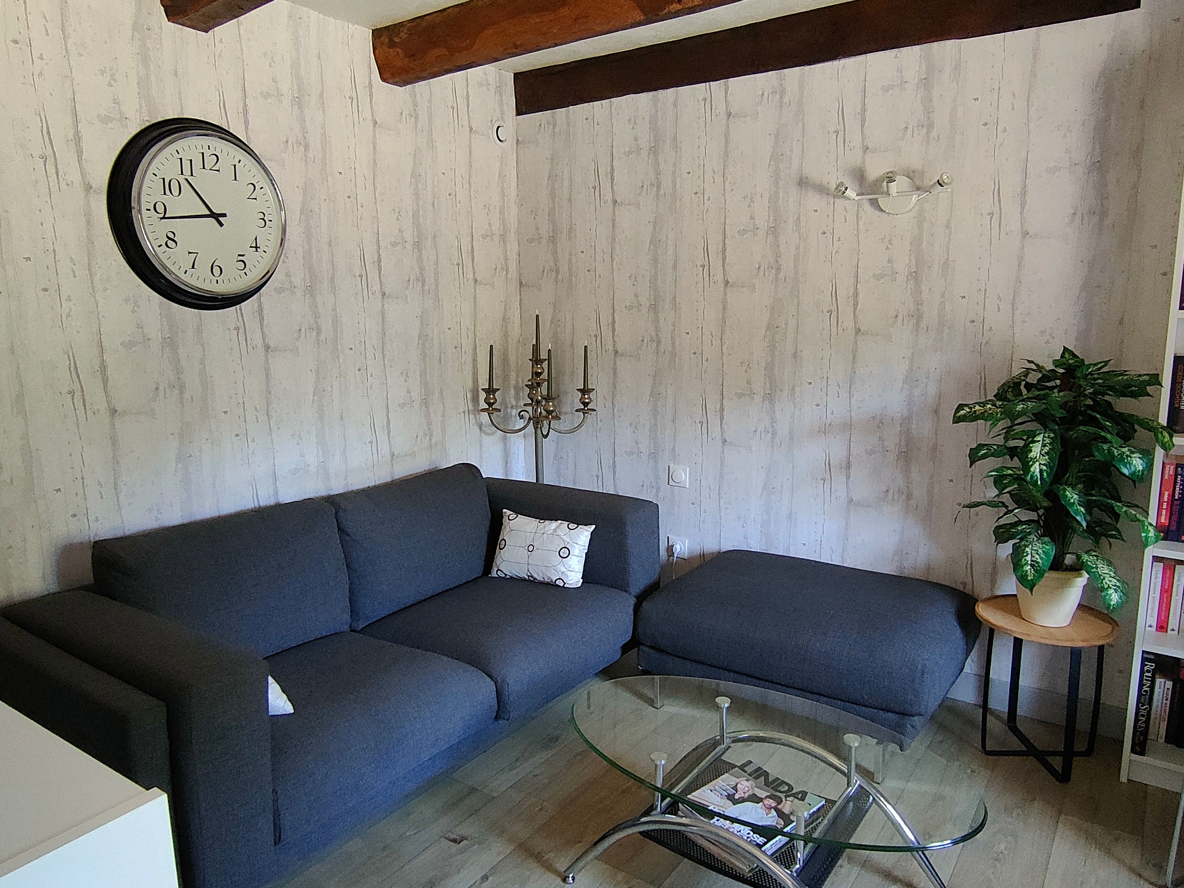 Een woonkamer met een hoekbank, een glazen salontafel, een wandklok, een kandelaarhouder en een potplant.