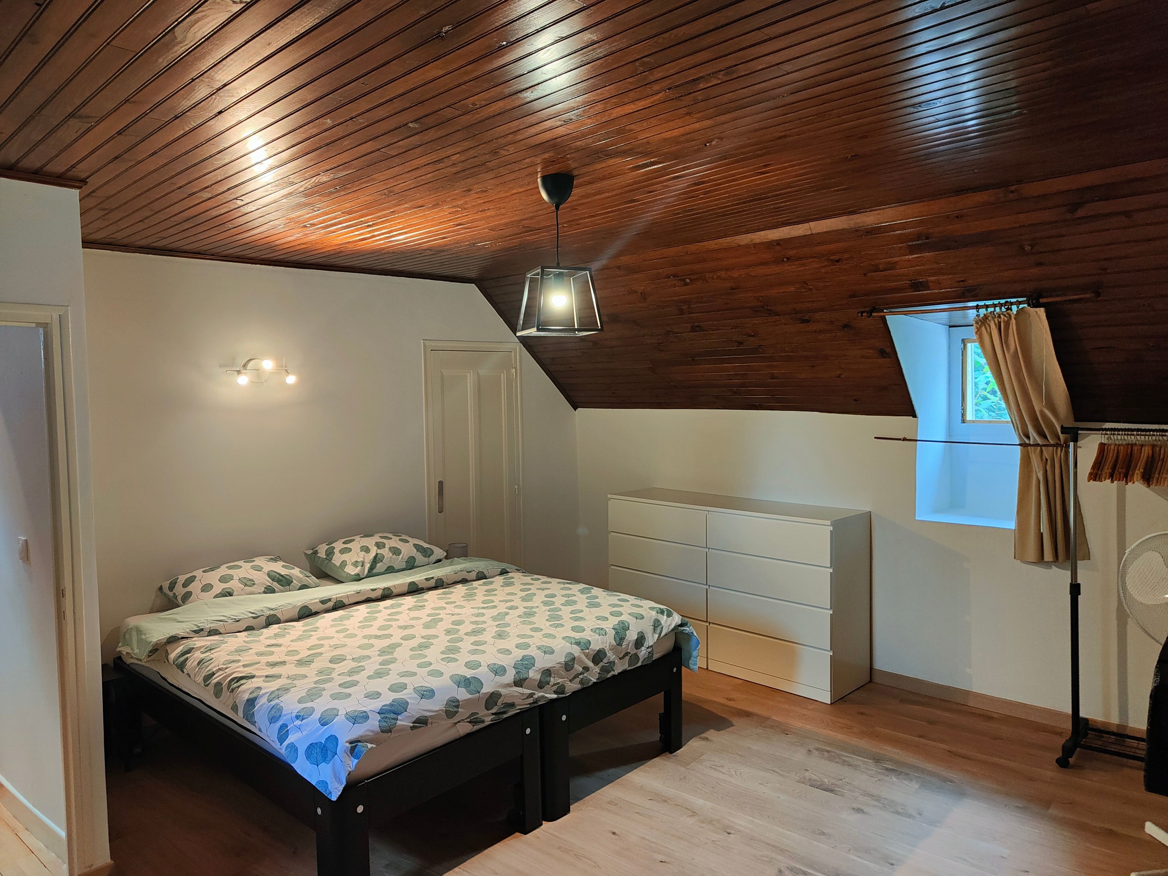 A bedroom with a sloped ceiling, a bed with patterned bedding, a bedside table, a dresser, and a window with curtains.