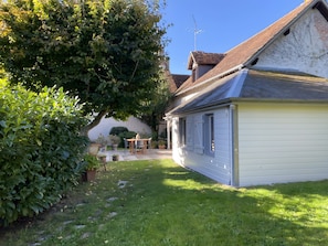 Una casa con techo inclinado, una chimenea visible y un jardín con césped y plantas en macetas.