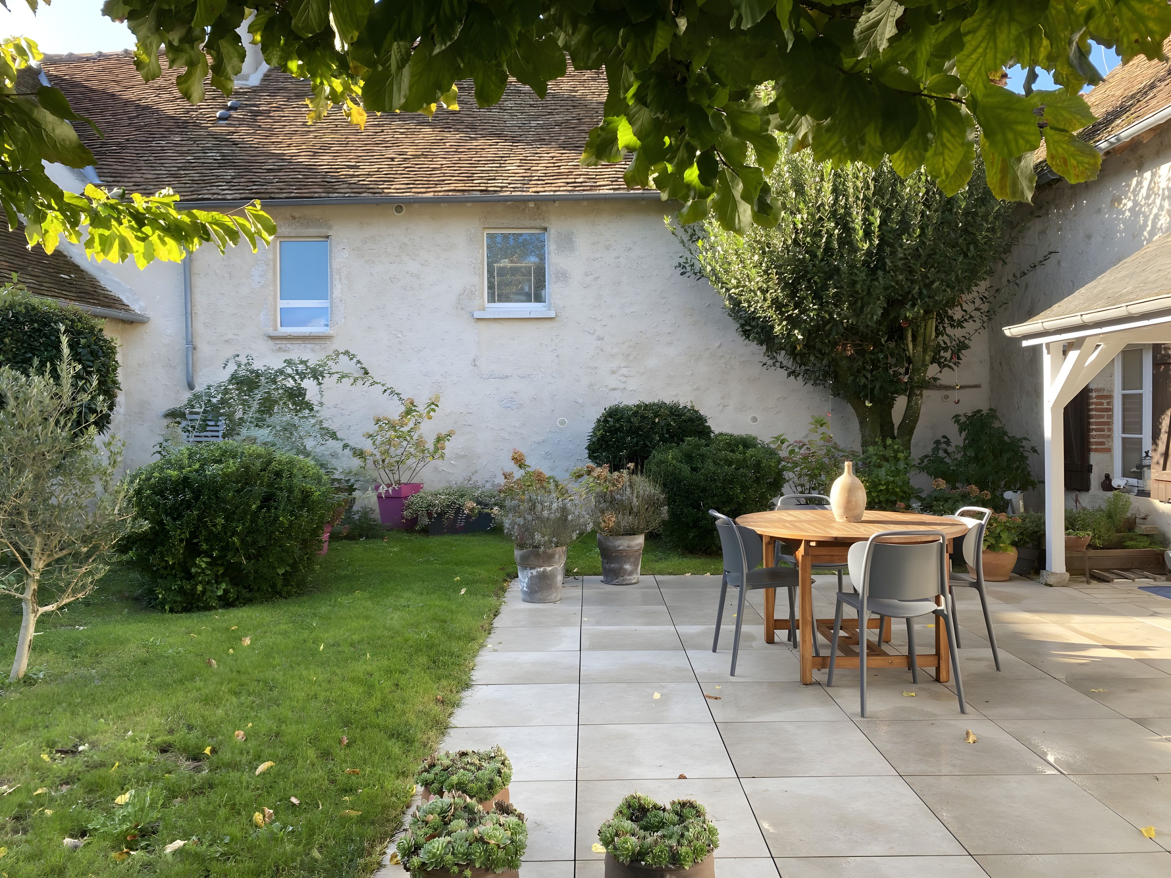 Une terrasse avec une table et des chaises, entourée de plantes en pot et d’une pelouse verdoyante.
