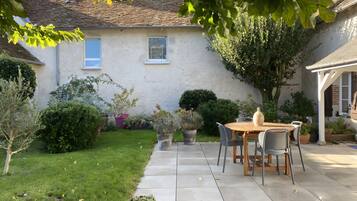 Une terrasse avec une table et des chaises, entourée de plantes en pot et d’une pelouse verdoyante.