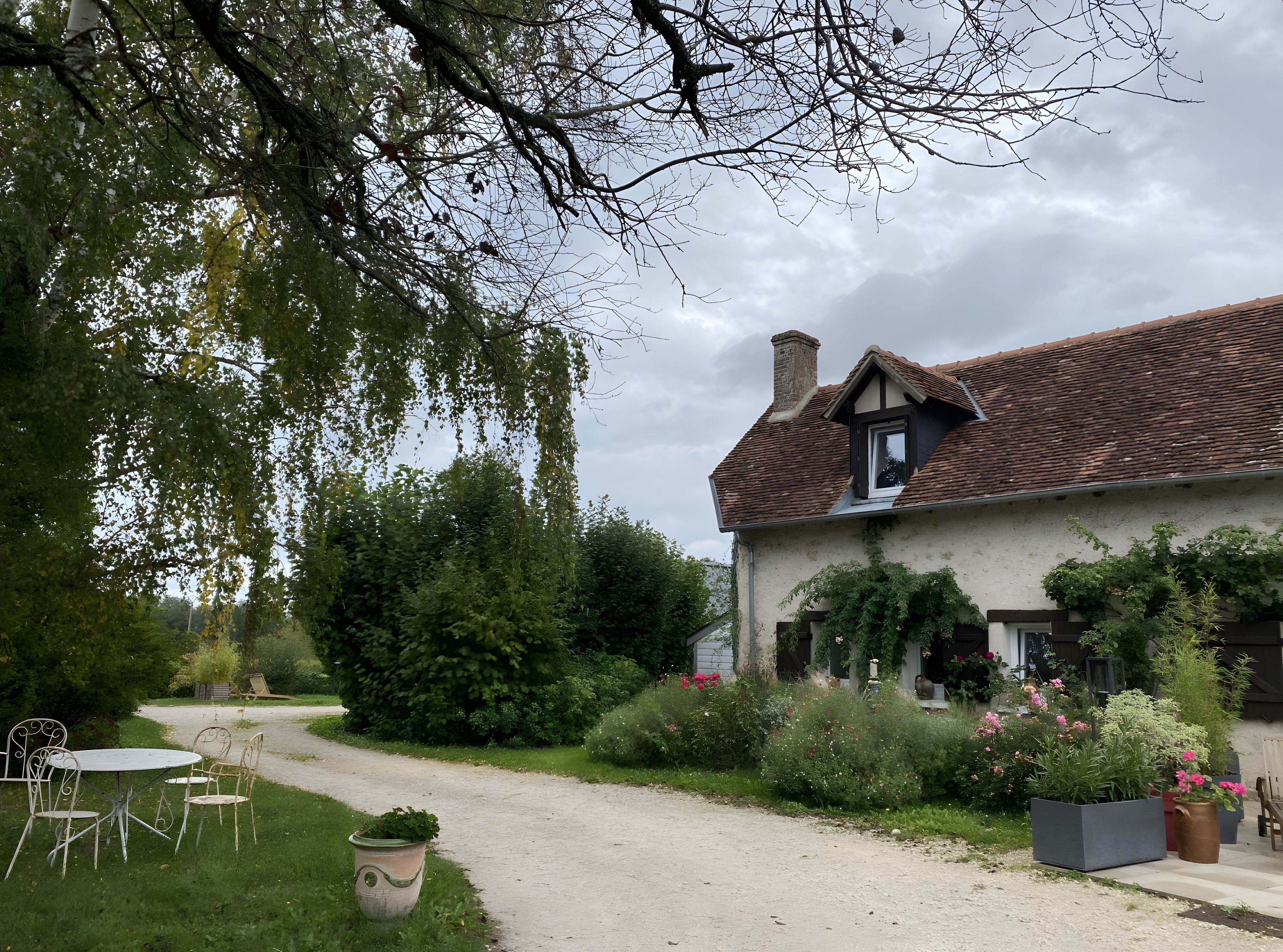 Une maison traditionnelle avec un toit en pente, entourée d’une végétation luxuriante et bordée d’un chemin de gravier menant à une porte blanche.