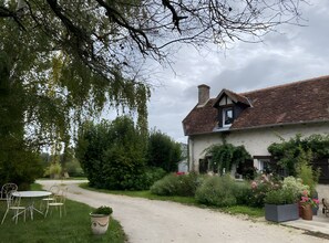 Ett traditionellt hus med ett sluttande tak, omgivet av frodig grönska och en grusgång som leder fram till en vit dörr.