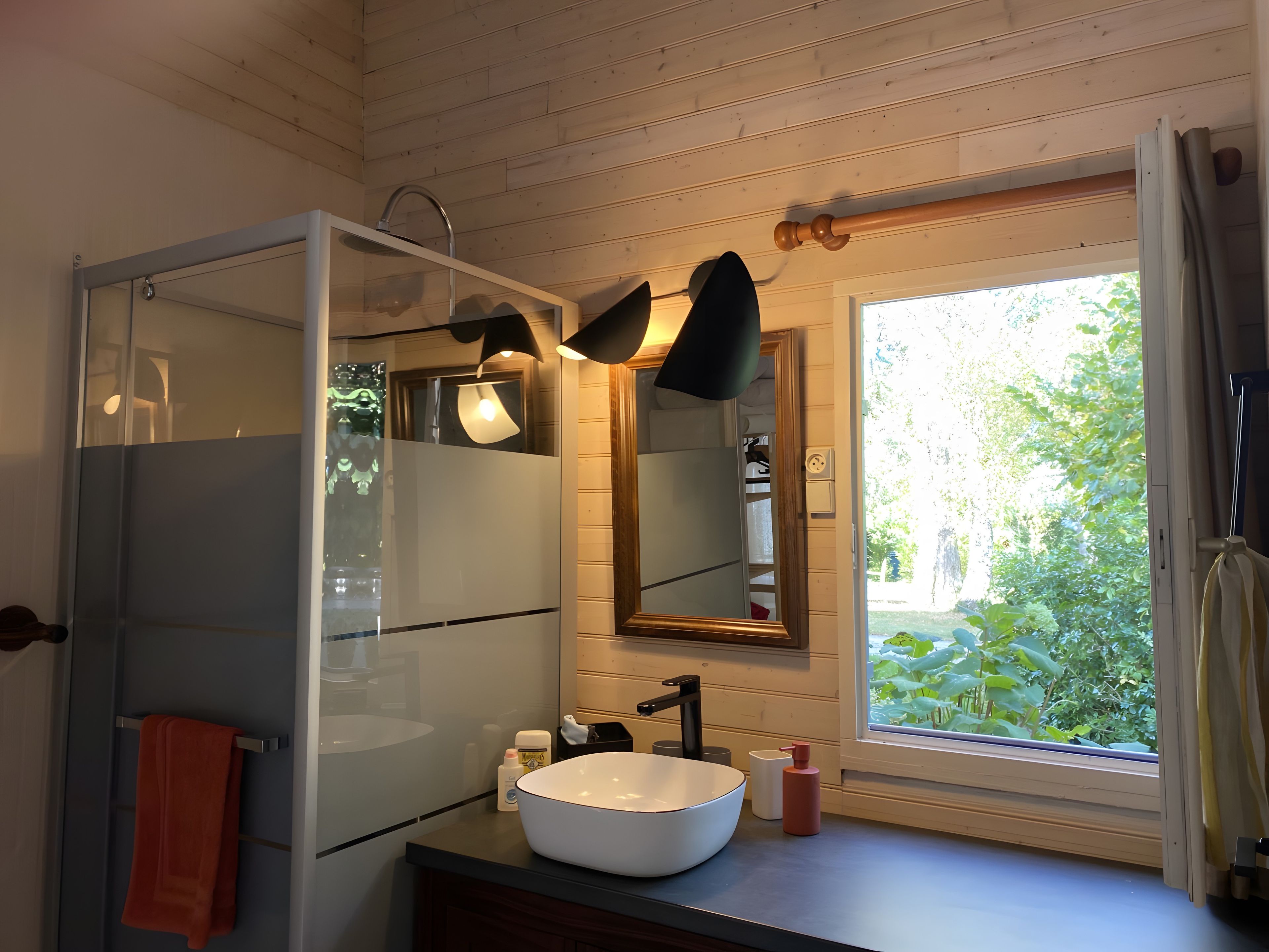 Un baño con ducha de vidrio, lavamanos blanco y ventana con vistas a la vegetación.