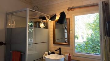 Un baño con ducha de vidrio, lavamanos blanco y ventana con vistas a la vegetación.