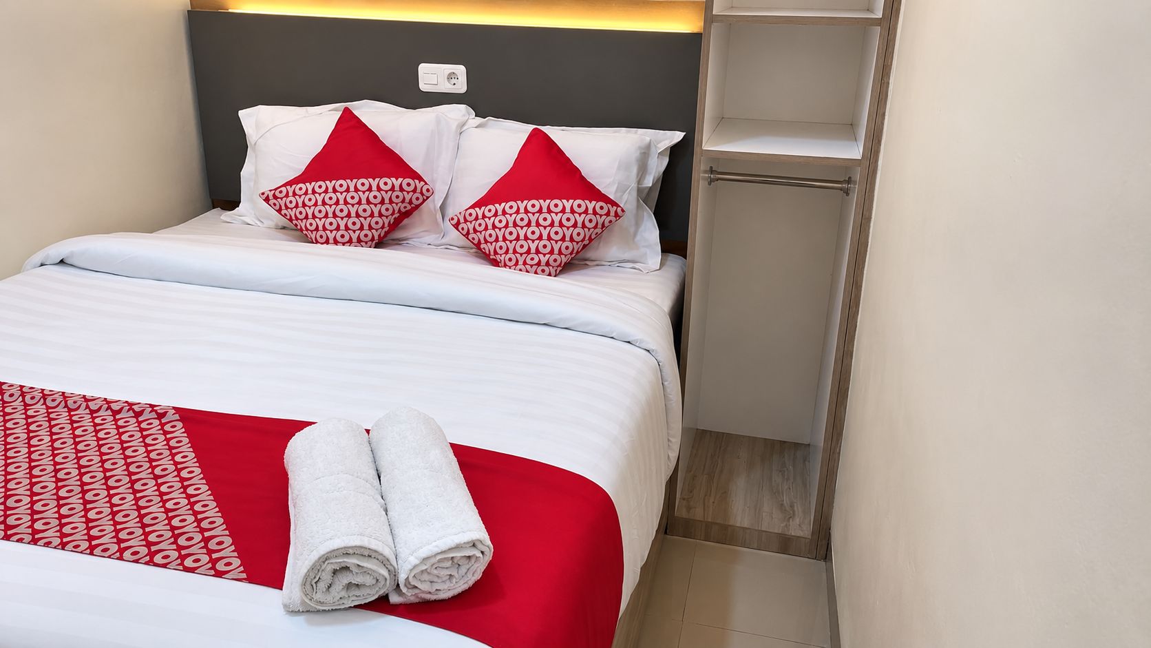 A neatly made bed with white bedding and red pillows, a pair of rolled towels, and a bedside shelf.