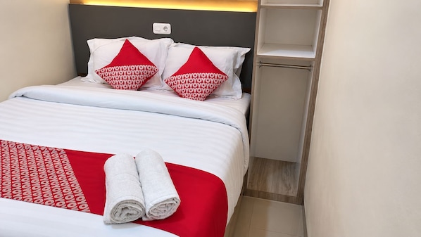 A neatly made bed with white bedding and red pillows, a pair of rolled towels, and a bedside shelf.