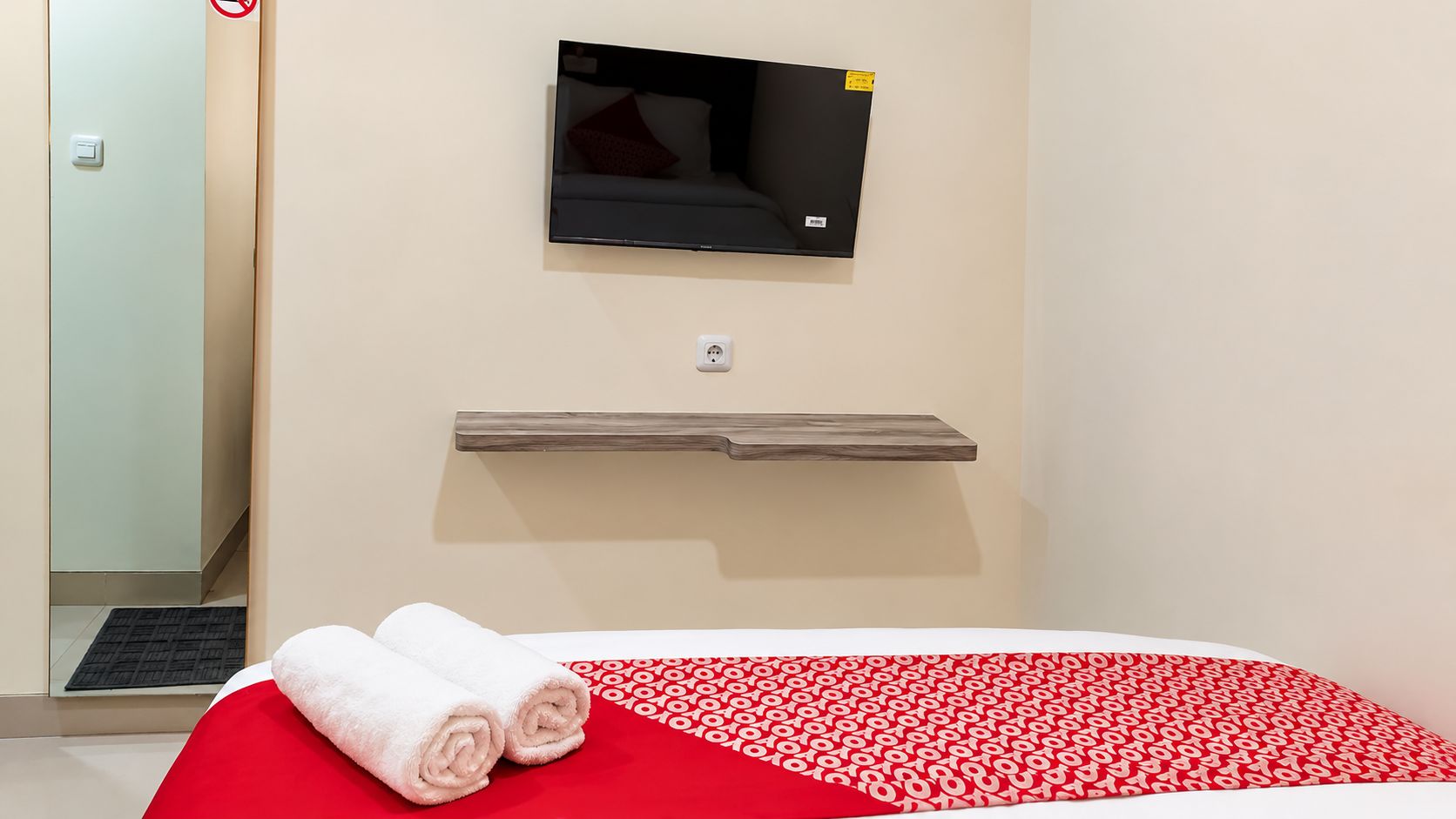 Una habitación de hotel con una cama, un televisor montado, un estante de madera y una puerta.