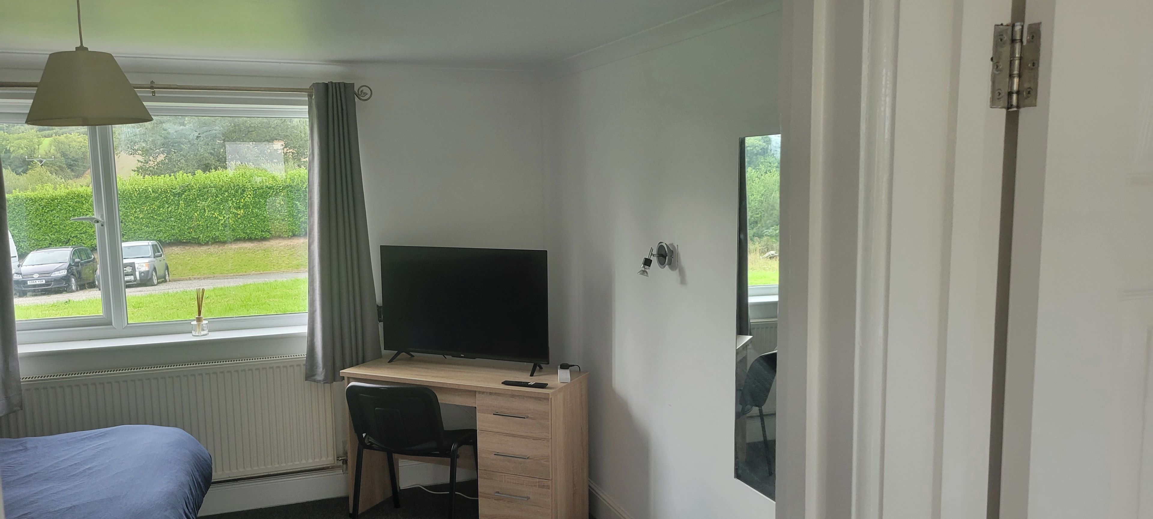 A bedroom with a bed, a desk with a computer, a chair, a window with curtains, and a view of the outside.