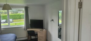 Een slaapkamer met een bed, een bureau met een computer, een stoel, een raam met gordijnen en uitzicht op het omliggende landschap.