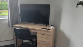 Una habitación con escritorio de madera, una silla negra, un televisor de pantalla plana y ventana con vista a coches estacionados y vegetación.