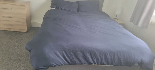 A bed with a grey mattress and a blue blanket in a room with a wooden dresser and a nightstand. - Apartment 2 Sleeps 2 With Enclosed Garden (Saltash)