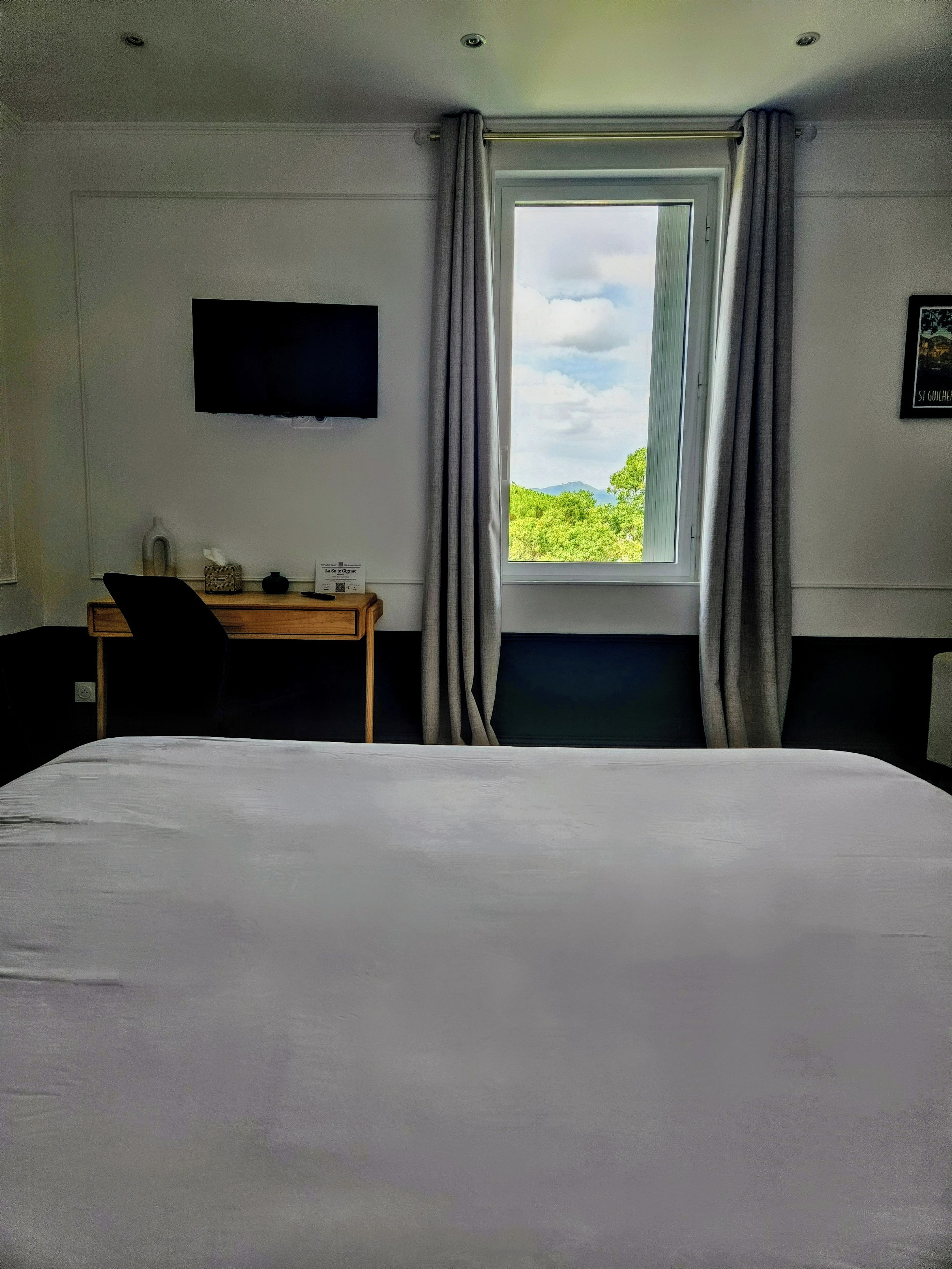 Una habitación de hotel con una cama, un escritorio, una silla, un televisor montado en la pared y una ventana con cortinas.