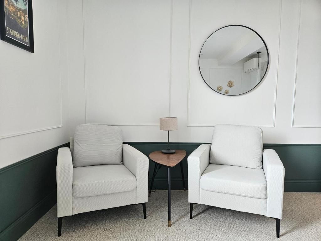 A room with two white armchairs, a small wooden table, a round mirror, and a framed picture on the wall.