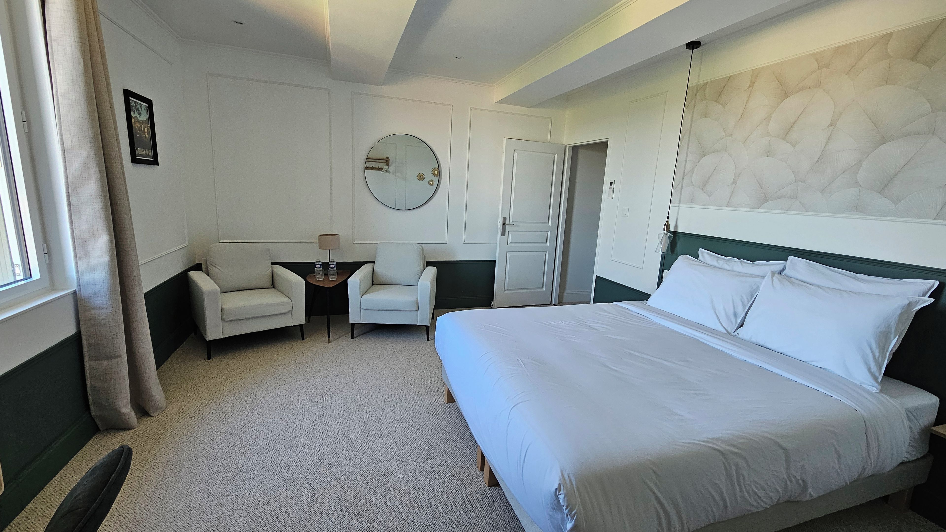 A hotel room with a large bed, two armchairs, a small table with a lamp, a round mirror, and a door.