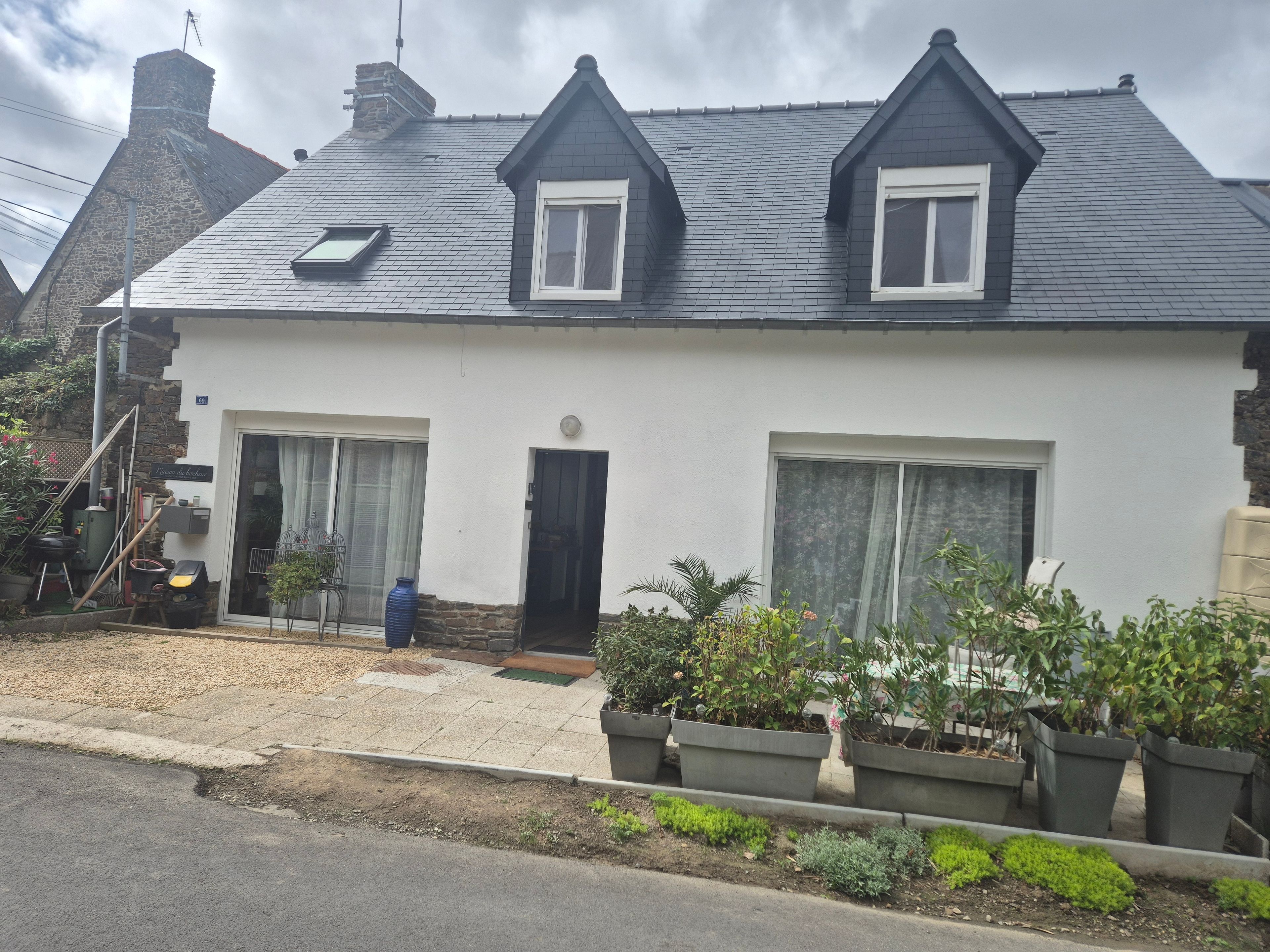 Une maison à deux étages, avec un toit sombre, des murs blancs et un petit porche agrémenté de plantes en pot.
