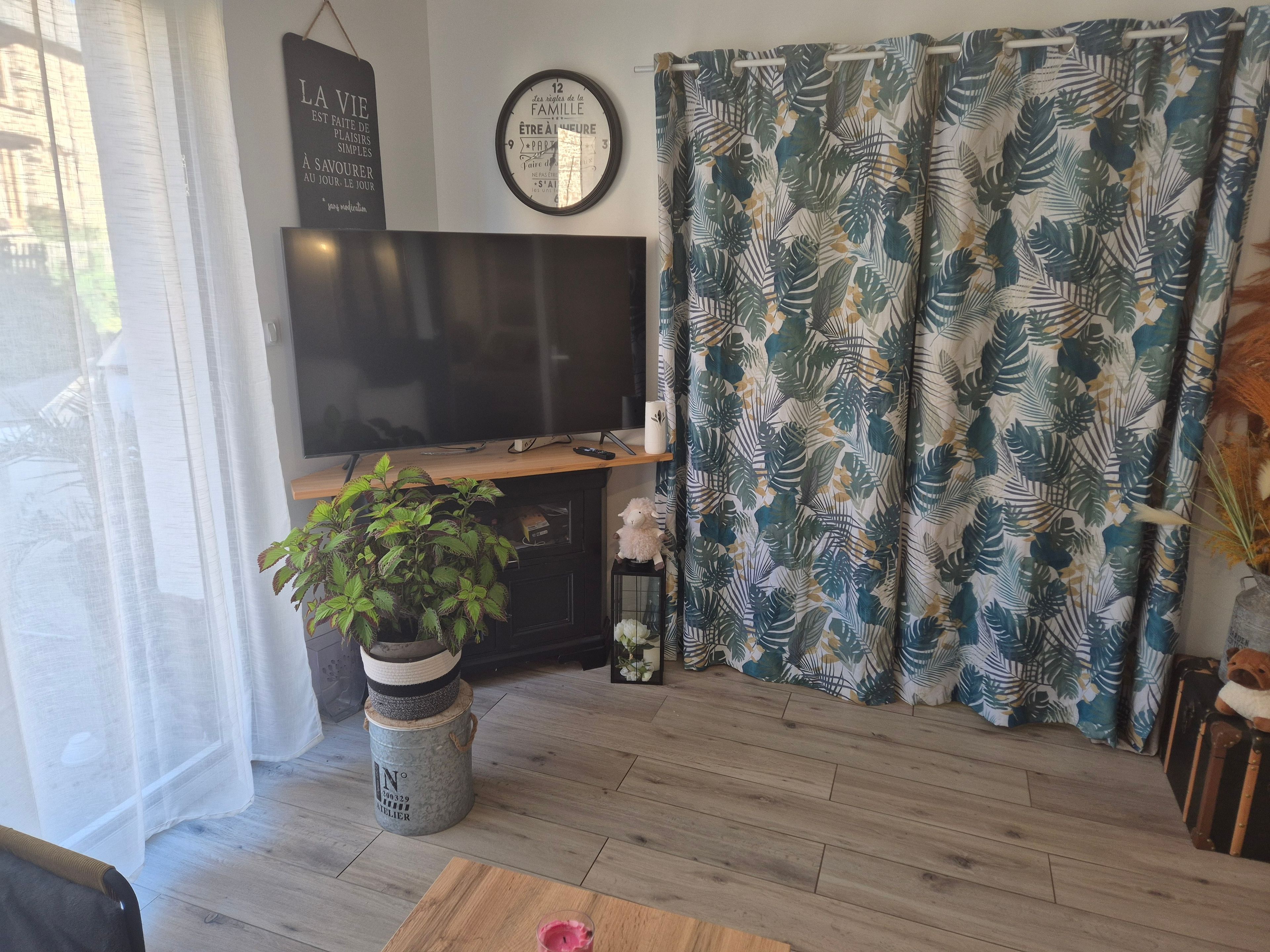 Ett vardagsrum med en tv, en krukväxt, en klocka och en gardin med ett tropiskt bladmotiv.