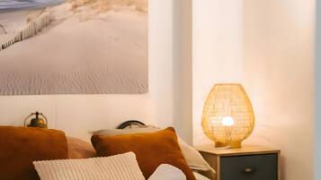 Une chambre avec une grande photographie d’une dune de sable accrochée au mur, un lit recouvert d’une couette blanche, deux oreillers marron, une serviette blanche, une table de chevet avec une lampe et une petite décoration représentant une carte du monde.