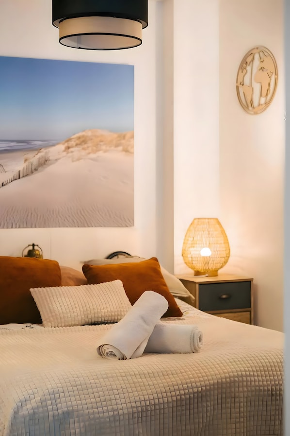 Ein Schlafzimmer mit einem großen Foto einer Sanddüne an der Wand, einem Bett mit weißer Decke, zwei braunen Kissen, einem weißen Handtuch, einem Nachttisch mit Lampe und einer kleinen Weltkarten-Dekoration.