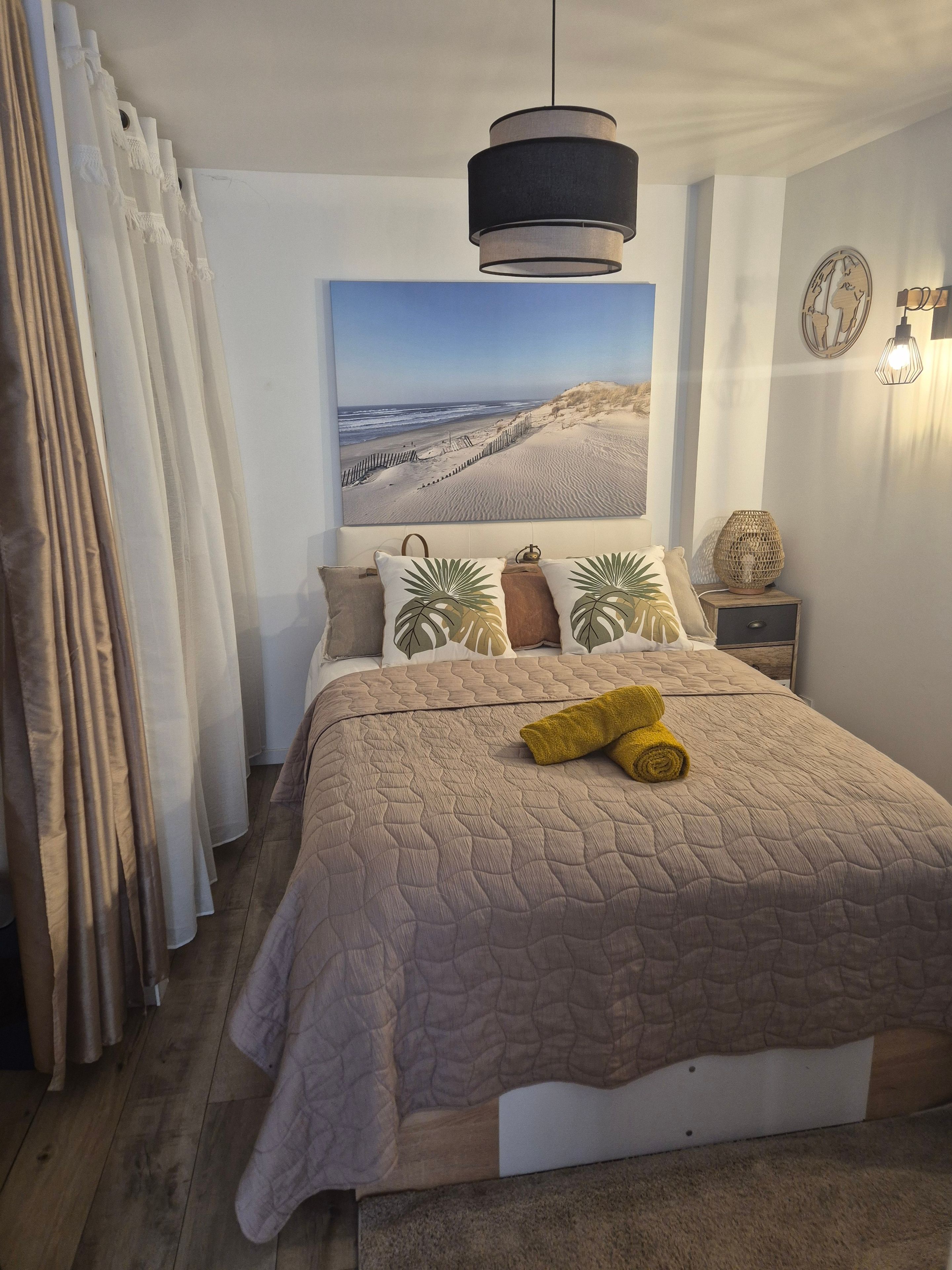 A bedroom with a large bed, a hanging light fixture, a wall-mounted lamp, and a beach-themed painting.
