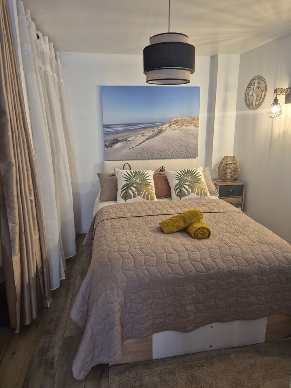 A bedroom with a large bed, a hanging light fixture, a wall-mounted lamp, and a beach-themed painting.