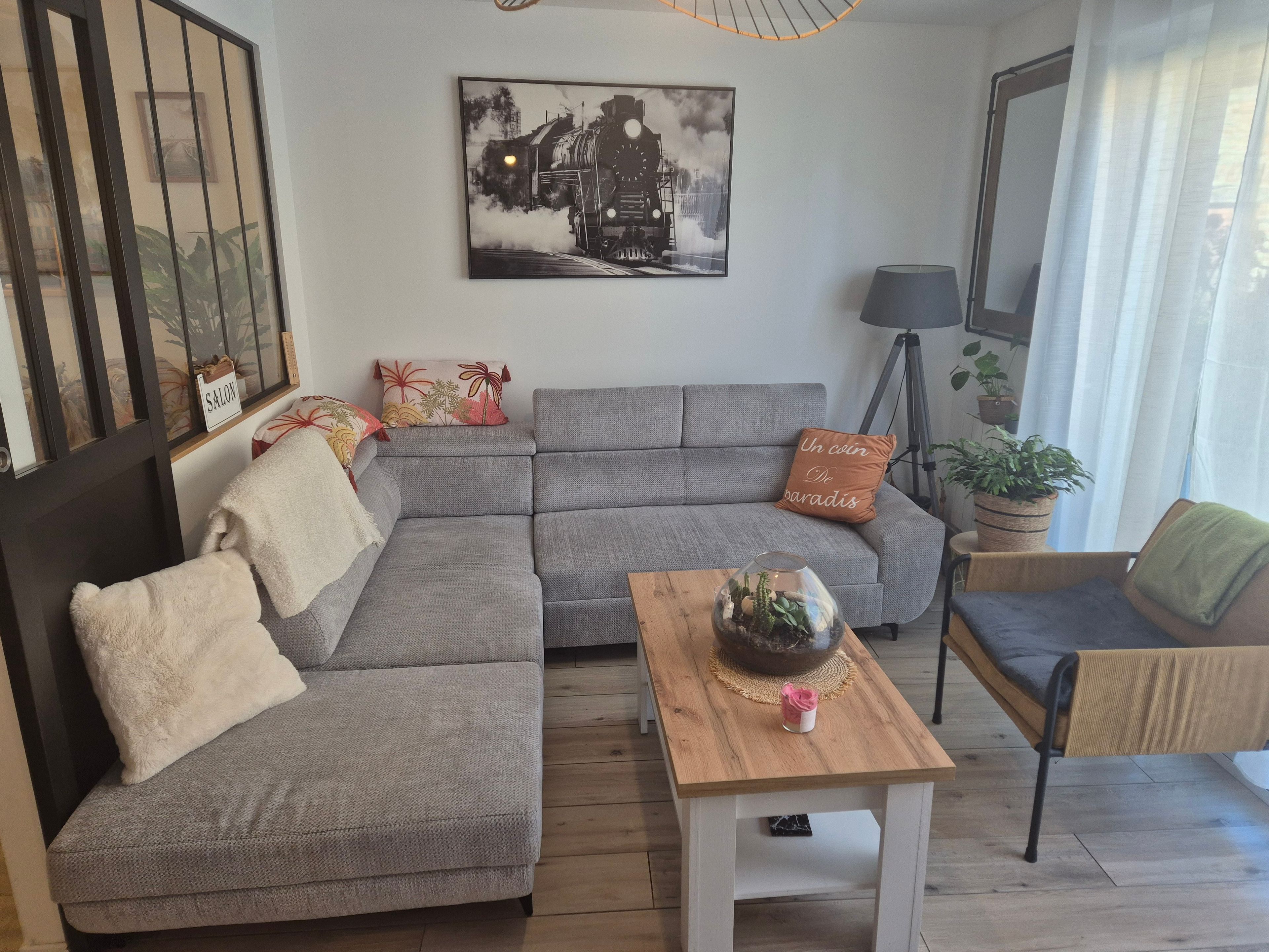 Un salon moderne avec un canapé d’angle gris, une table basse en bois et un tableau en noir et blanc représentant un train, accroché au mur.