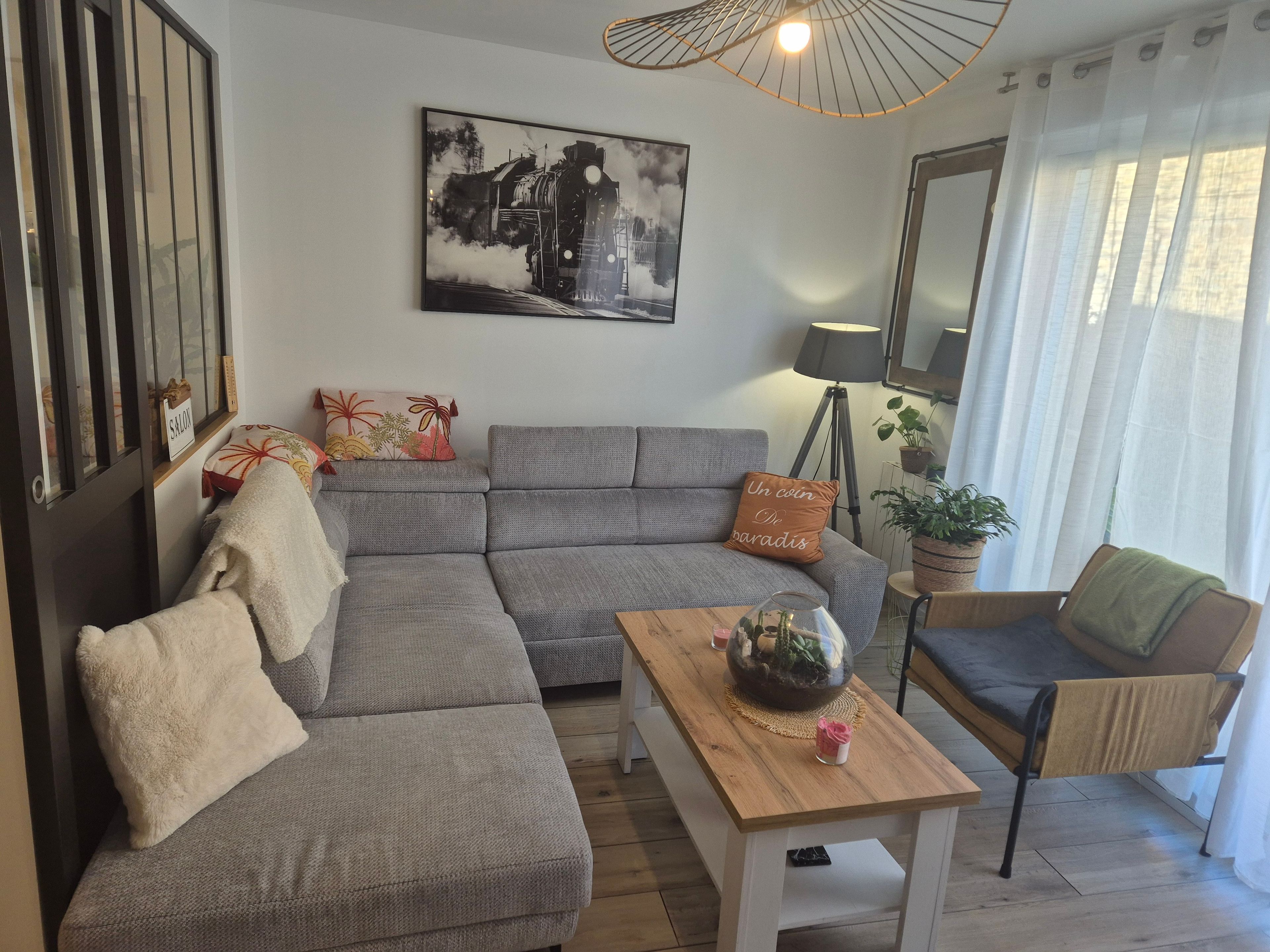Un salon moderne avec un canapé d’angle gris, une table basse en bois, un lampadaire et un tableau accroché au mur.