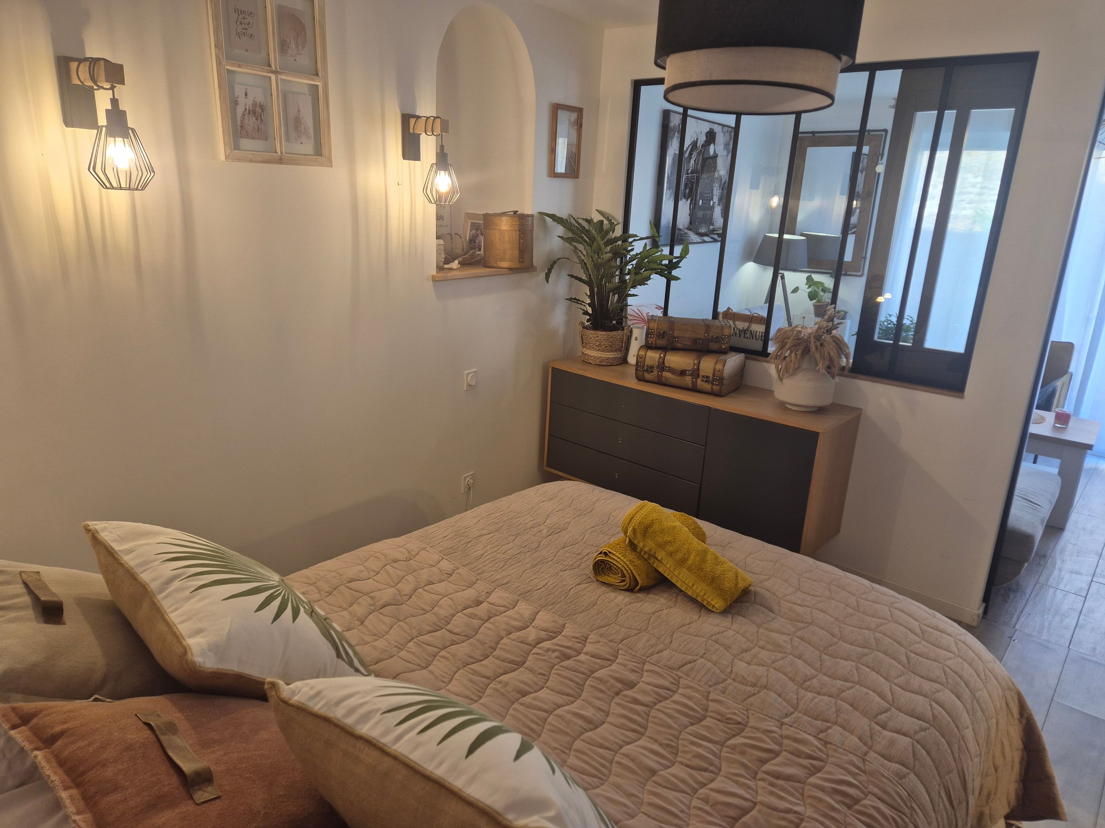 Une chambre à coucher avec un lit, une commode, des lampes fixées au mur, une fenêtre et une plante en pot.