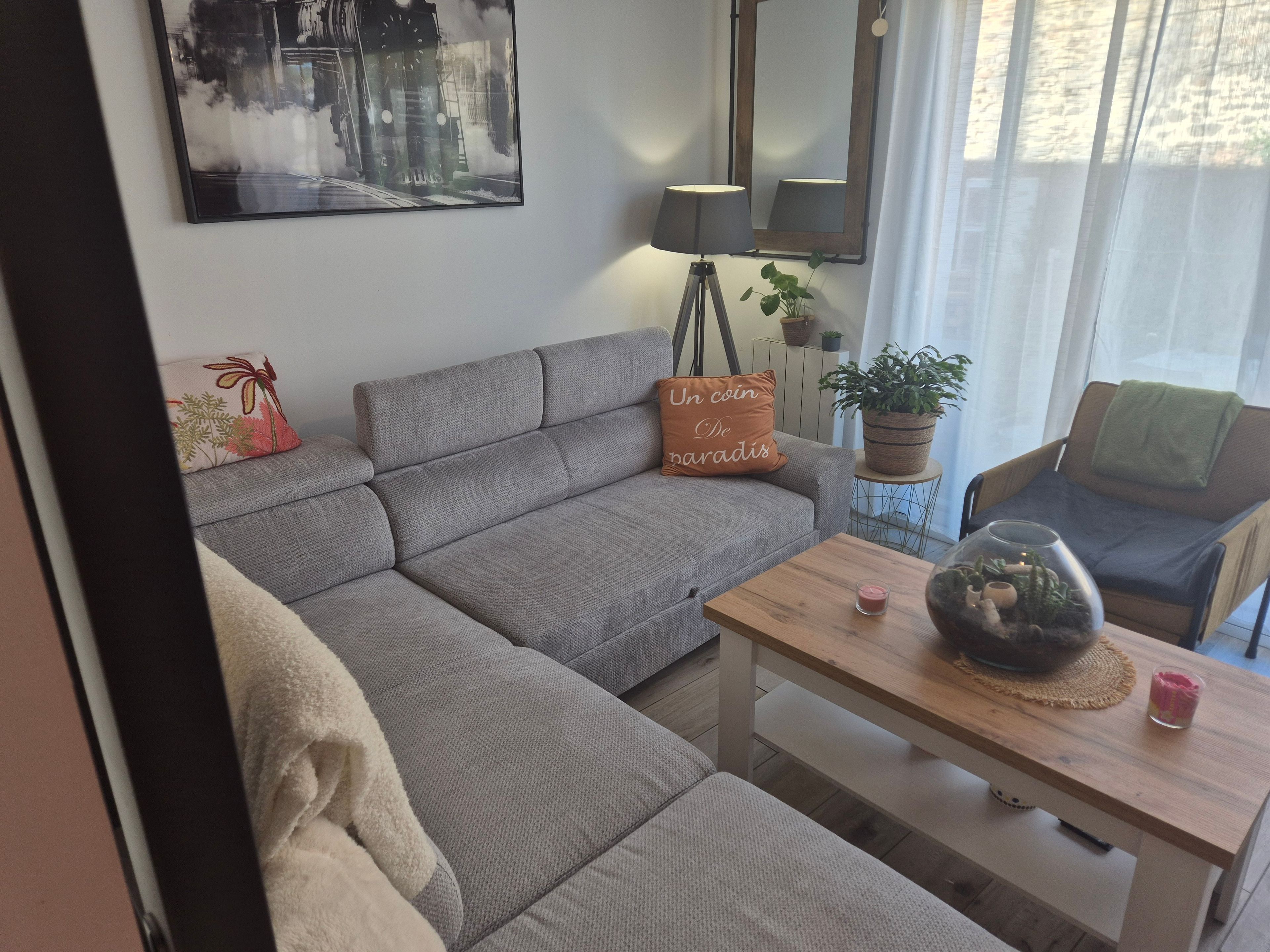 Ett modernt vardagsrum med en grå hörnsoffa, ett litet träbord, en golvlampa, en krukväxt och en tavla som hänger på väggen.