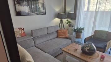 Ett modernt vardagsrum med en grå hörnsoffa, ett litet träbord, en golvlampa, en krukväxt och en tavla som hänger på väggen.