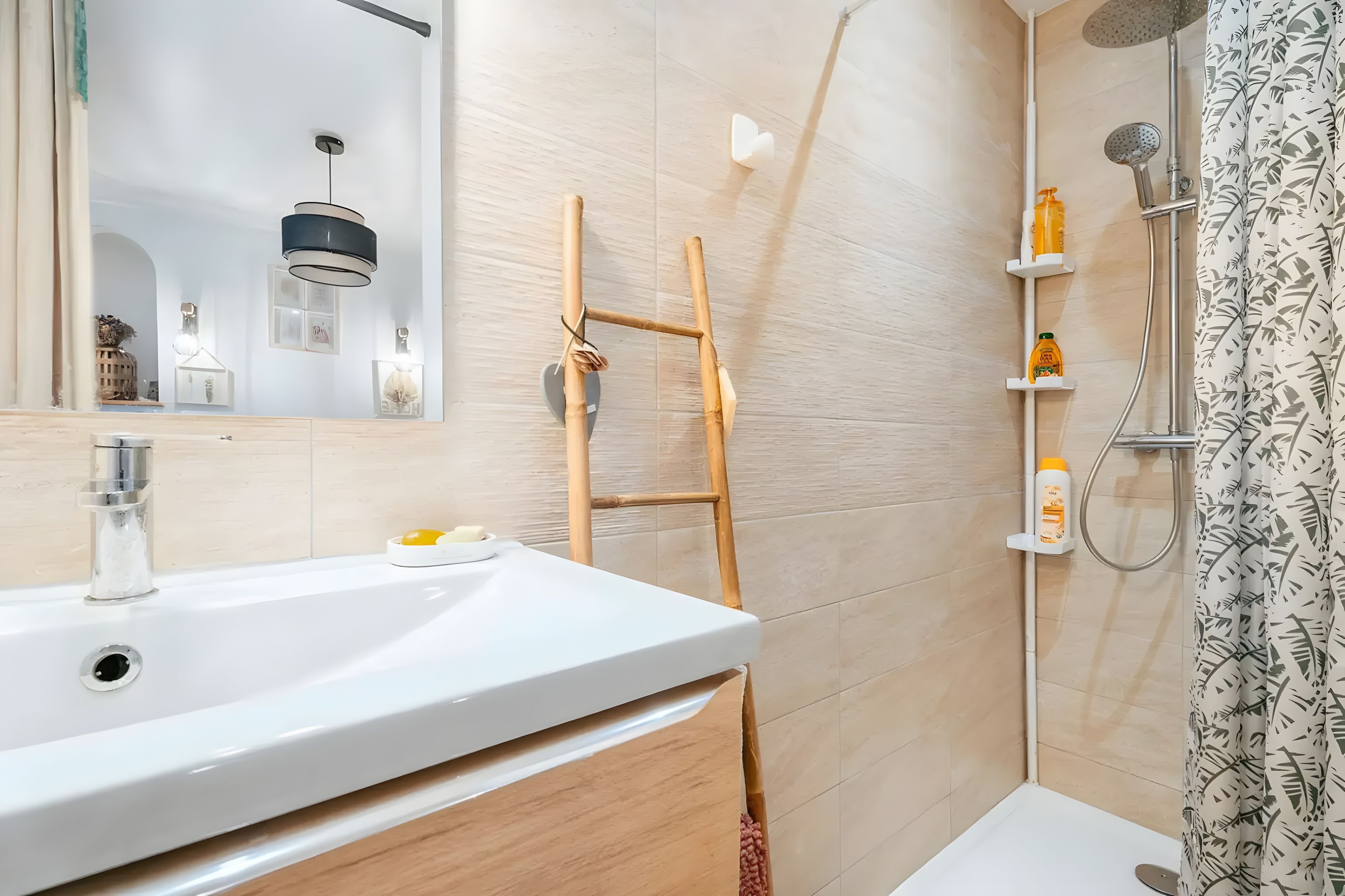 Een badkamer met een witte wastafel, een spiegel, een douche met douchehouder en een douchegordijn met een bladmotief.