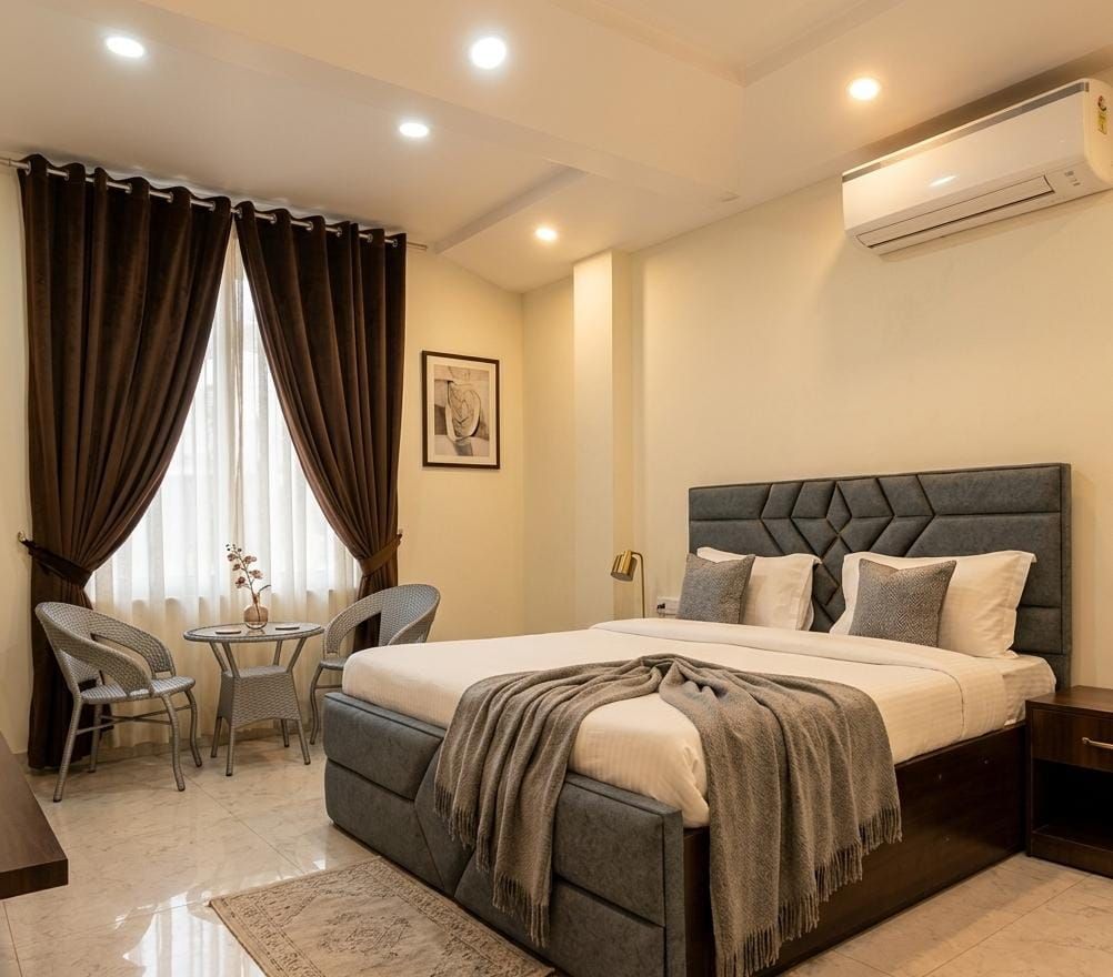 A bedroom with a bed, bedside tables, a chair, a small table, and an air conditioning unit.
