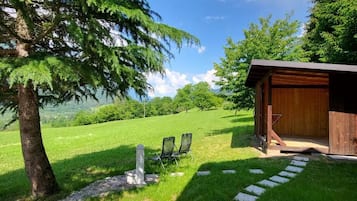 A wooden cabin with a stone pathway leading to two chairs, surrounded by lush greenery and a clear sky.