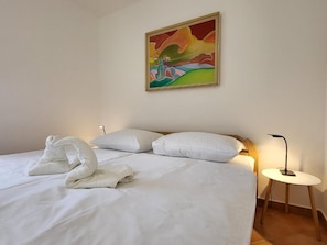 A hotel room with a bed, white pillows, a painting on the wall, and a bedside table with a lamp.