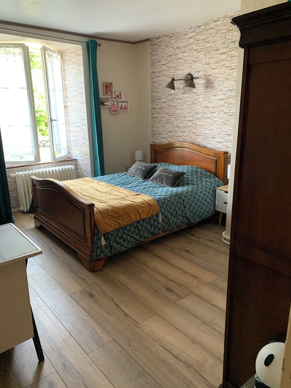 A bedroom with a wooden bed, a radiator, a window with curtains, a stone wall, and a small bedside table.