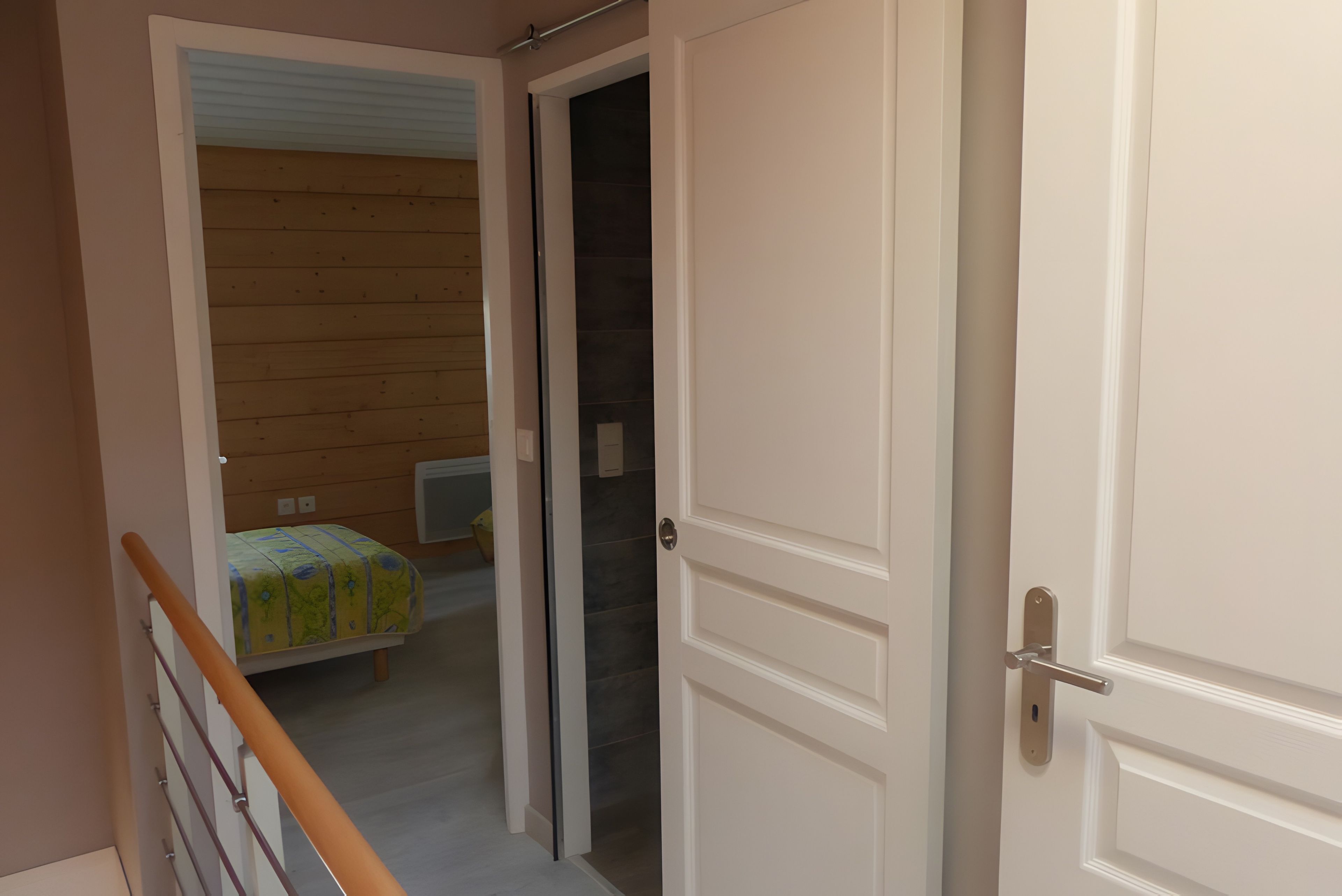 Un couloir au plancher de bois mène à une pièce où se trouvent un lit et une fenêtre.