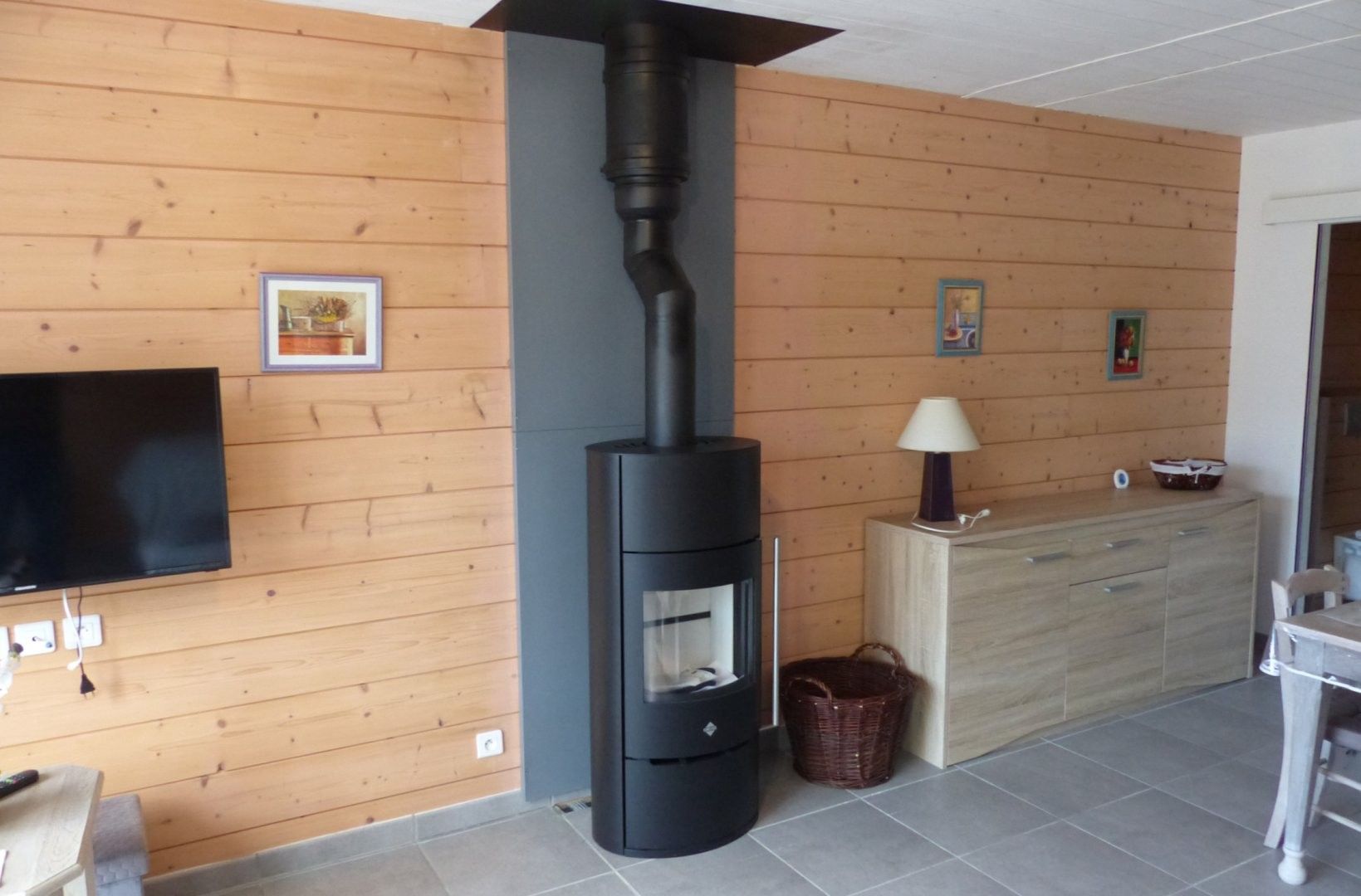 Een kamer met een houtkachel, een flatscreen televisie aan de muur, een houten kast en een rieten mand op de vloer.