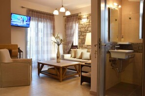 Ein Wohnzimmer mit Sofa, Couchtisch und Fernseher. Eine Steinwand und ein Badezimmer mit Waschbecken und Spiegel sind ebenfalls vorhanden.