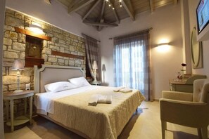 A hotel room with a stone wall, a bed with white linens, a wooden headboard, a bedside table with a lamp, a chair, and a large window with curtains. - Liokrina Villa Elia - Livadakia Poolside Gem (Λιβαδάκια)