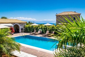 A swimming pool with lounge chairs and umbrellas, surrounded by greenery and a stone building. - Liokrina Villa Elia - Livadakia Poolside Gem (Λιβαδάκια)