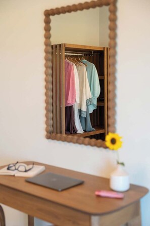 A mirror reflecting a clothing rack with various garments, a desk with a laptop, glasses, and a pink pen, and a vase with a yellow flower. - Liokrina Suite 03 - Livadakia Poolside Getaway! (Livadakia)