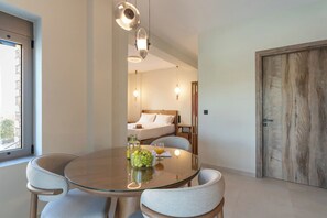 A modern hotel room with a round glass table, two chairs, a bed with white linens, a wooden door, and a window with a view. - Liokrina Suite 03 - Livadakia Poolside Getaway! (Livadakia)