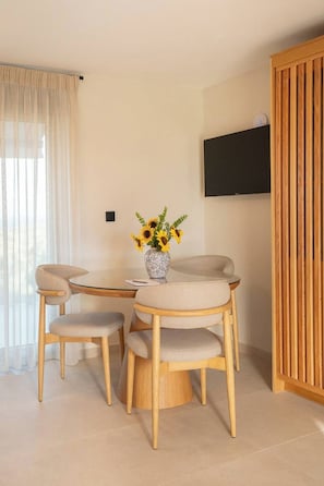 A dining area with a round table, four chairs, a vase with sunflowers, a television on the wall, and a wooden panel on the right. - Liokrina Suite 05 - Livadakia Poolside Getaway! (Livadakia)