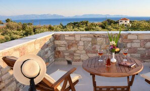 Un muro de piedra con vistas al mar y a las montañas, una mesa de madera con bebidas y un libro, y un sombrero de paja sobre una silla.