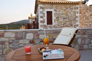 A stone building with a tiled roof, a table with a drink, a chair, and a book. - Liokrina Villa Hellios - Livadakia Poolside Gem (Λιβαδάκια)