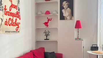 A living room with a red sofa, a small table, a lamp, and a shelf with decorative items.