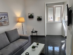 Una sala moderna con un sofá gris, una mesa de centro blanca y una lámpara. Hay dos mesitas auxiliares con adornos. Un cuadro está en la pared, y se ve una ventana con persianas.