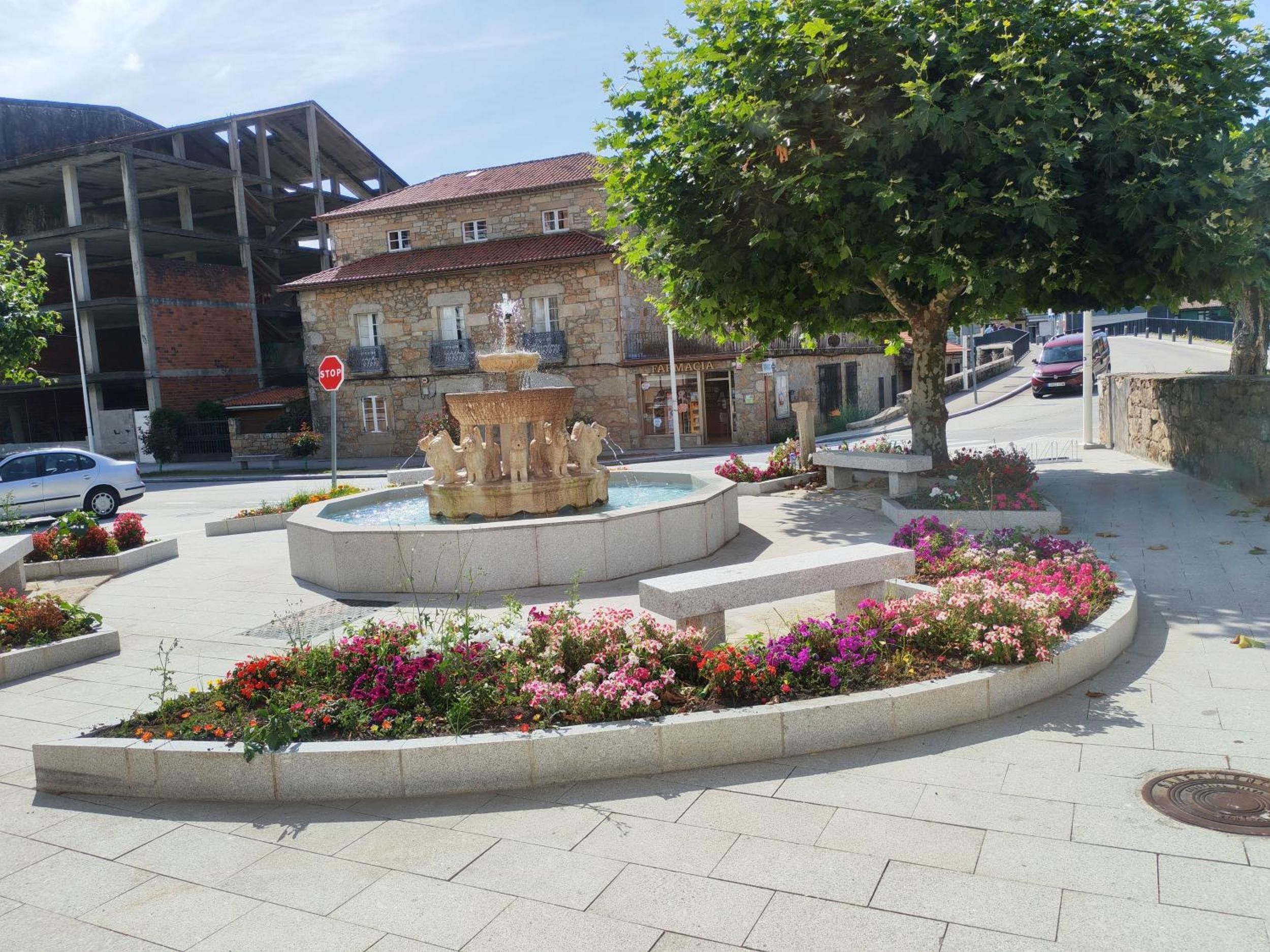 Una plaza pública con una fuente central, rodeada de macizos de flores y un edificio de piedra al fondo.