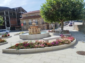 Una plaza pública con una fuente central, rodeada de macizos de flores y un edificio de piedra al fondo.