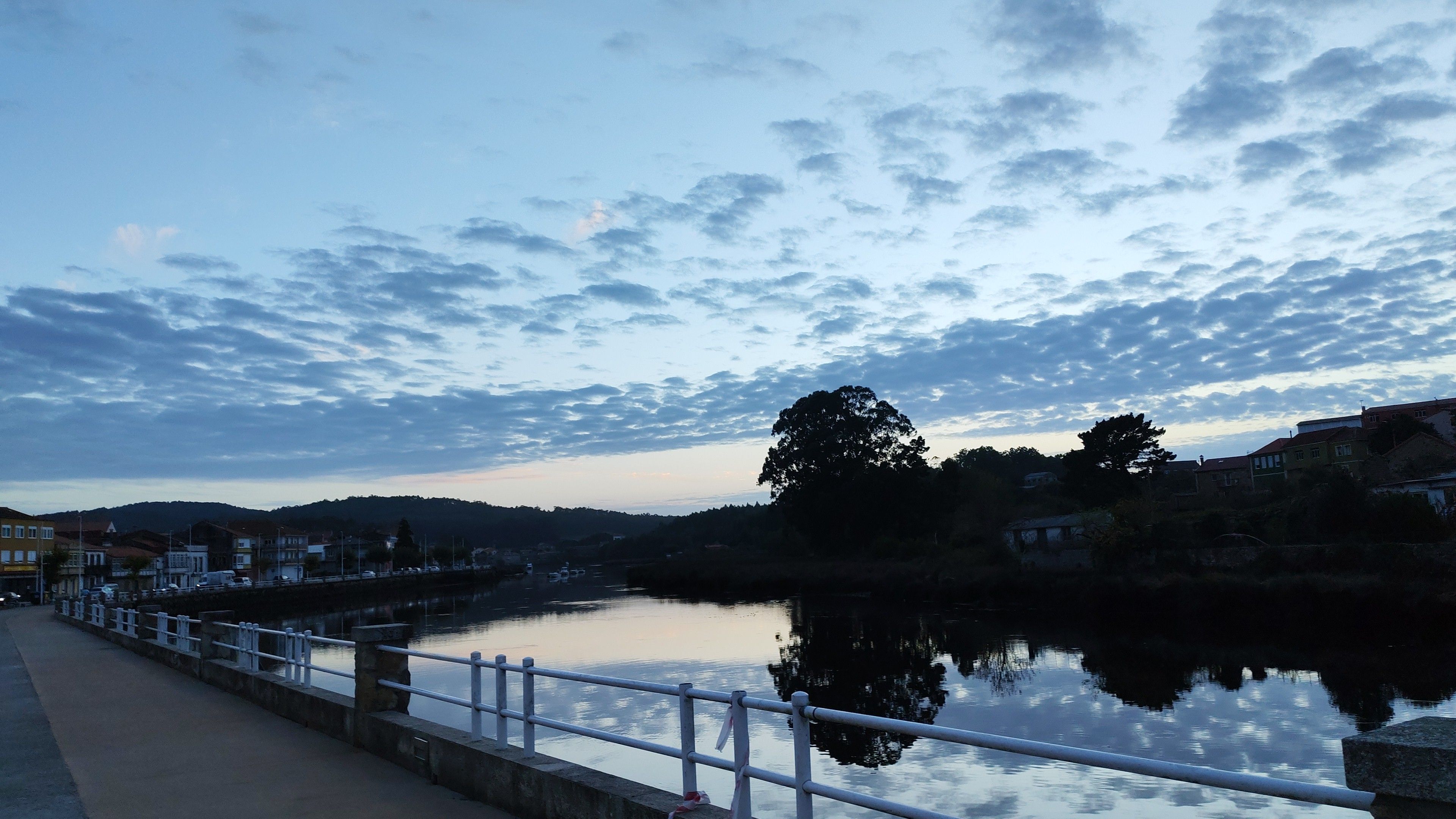 A riverside view with a calm water body, a bridge, and a row of buildings along the riverbank.